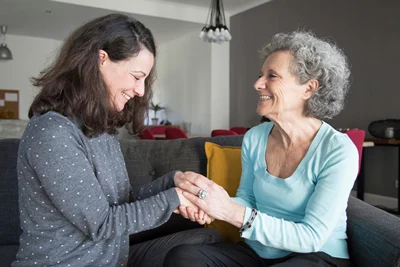 Two women of different ages sit on a couch in a living room, smiling and holding hands in a warm, supportive moment.