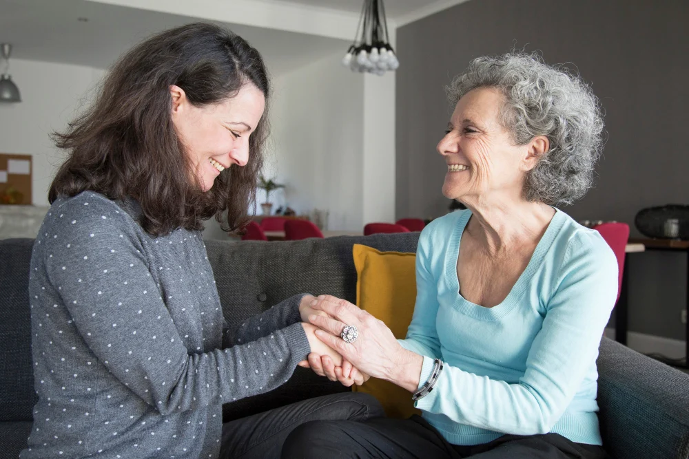 Two women sit on a couch, smiling warmly while clasping hands in a moment of friendship and support.