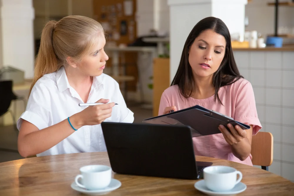 Two women reviewing a clipboard beside a laptop at a table