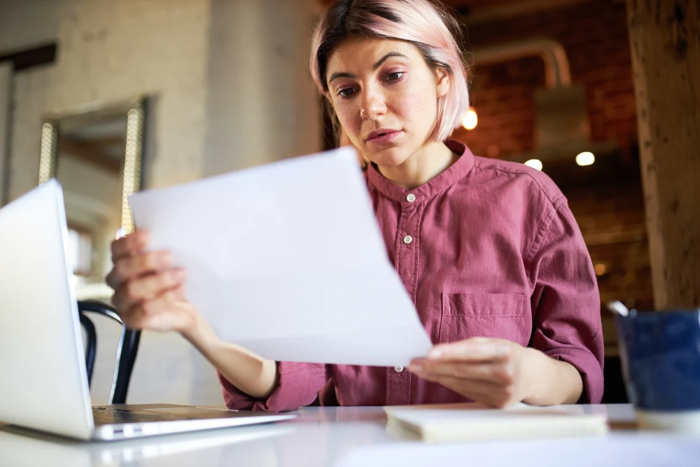 Woman carefully reviewing NDIS service agreement beside laptop Woman carefully reviewing NDIS service agreement beside laptop